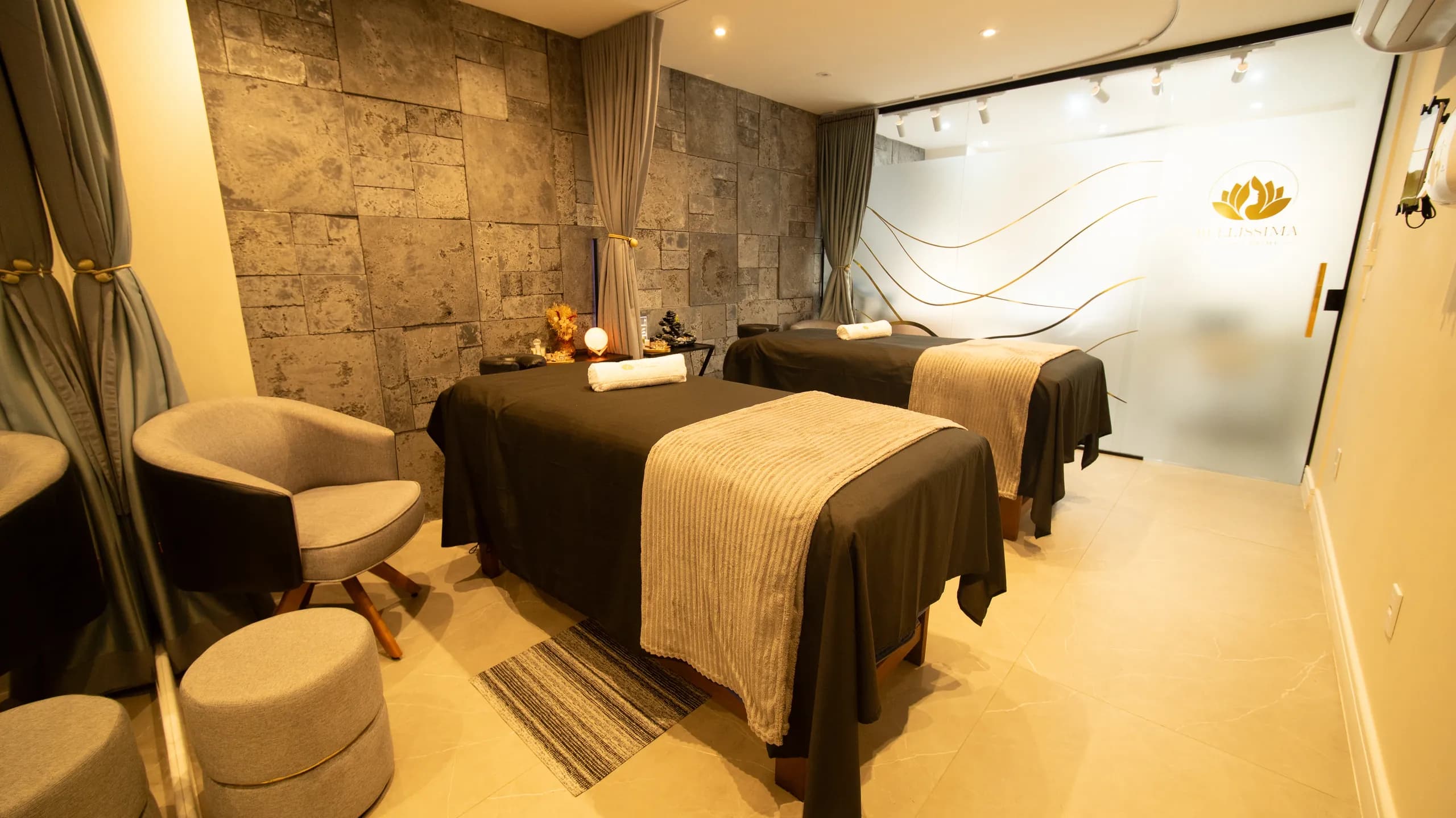 Sala de massagem com duas macas, parede de pedra e cortinas