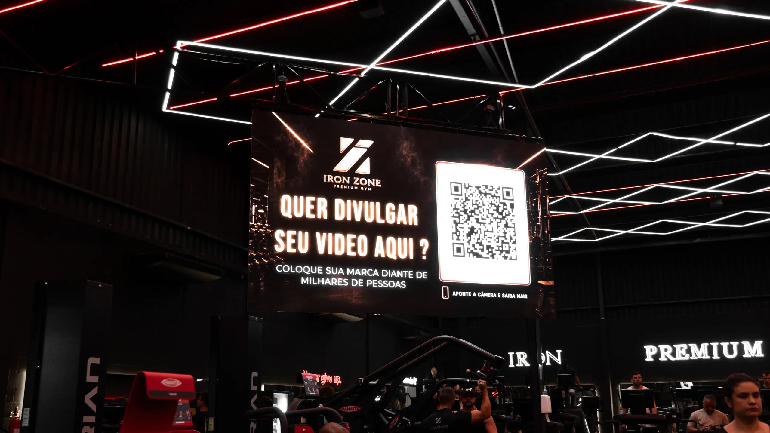 Telão central do salão da Iron Zone exibindo a chamada "Quer divulgar seu vídeo aqui?" com QR code, sob o teto de LED